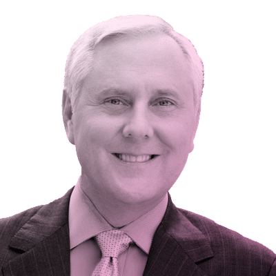 A middle-aged man with light hair in a suit and tie is smiling at the camera. The image has a pinkish tint and a plain white background.